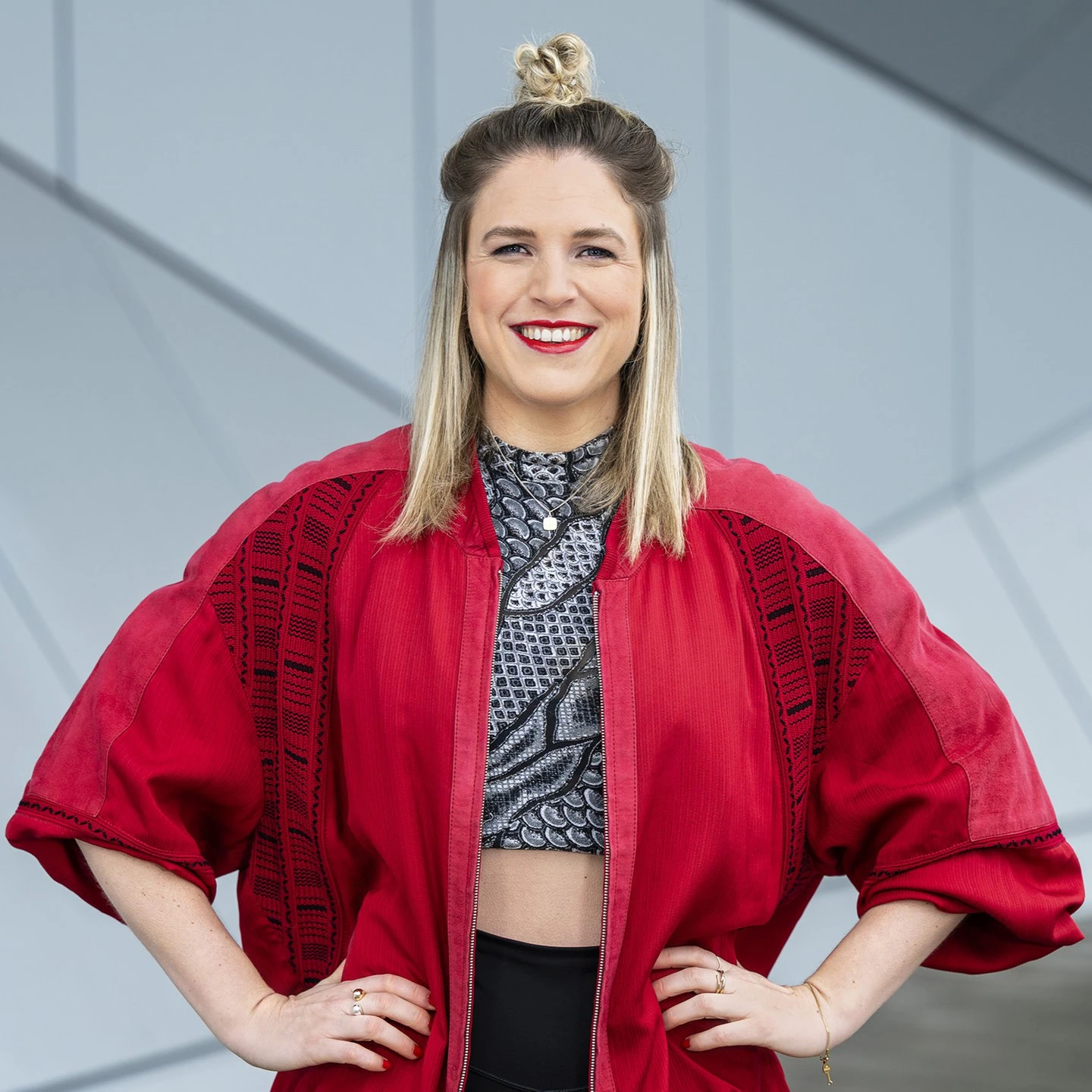 Lächelnde Person Dani Woytewicz in roter Jacke vor moderner grauer Architektur, selbstbewusst in die Kamera blickend.