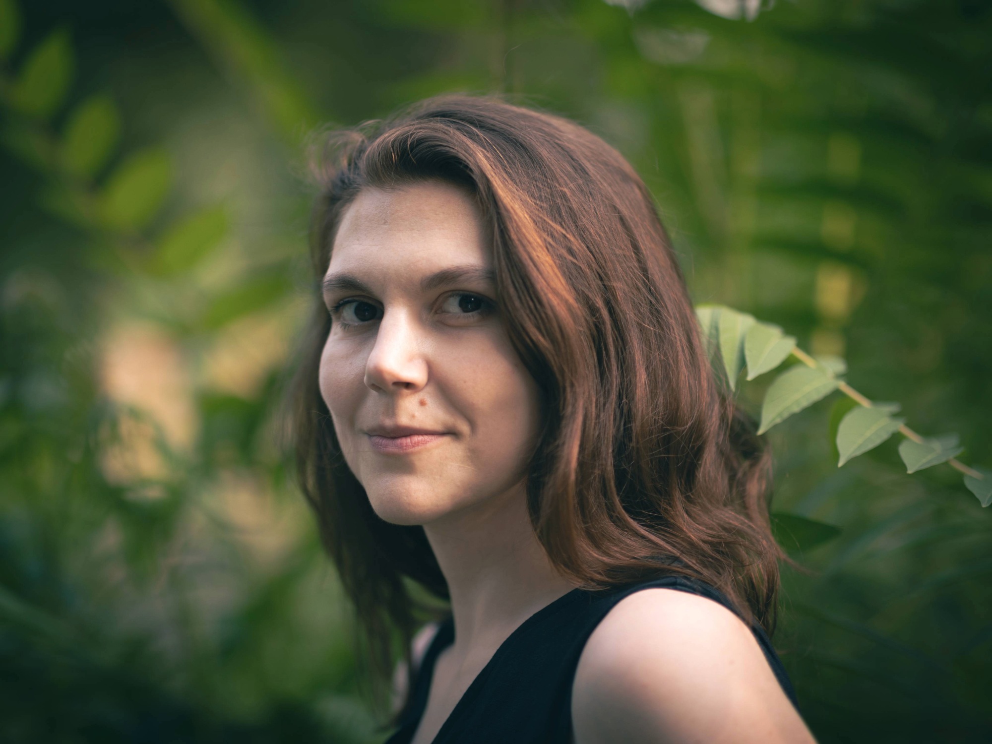 Ana Wetherall-Grujic: Dunkelhaarige Frau schaut im Viertelprofil in die Kamera. Sie trägt ein schwarzes Kleid. Im Hintergrund sind grüne Pflanzen zu sehen. 
