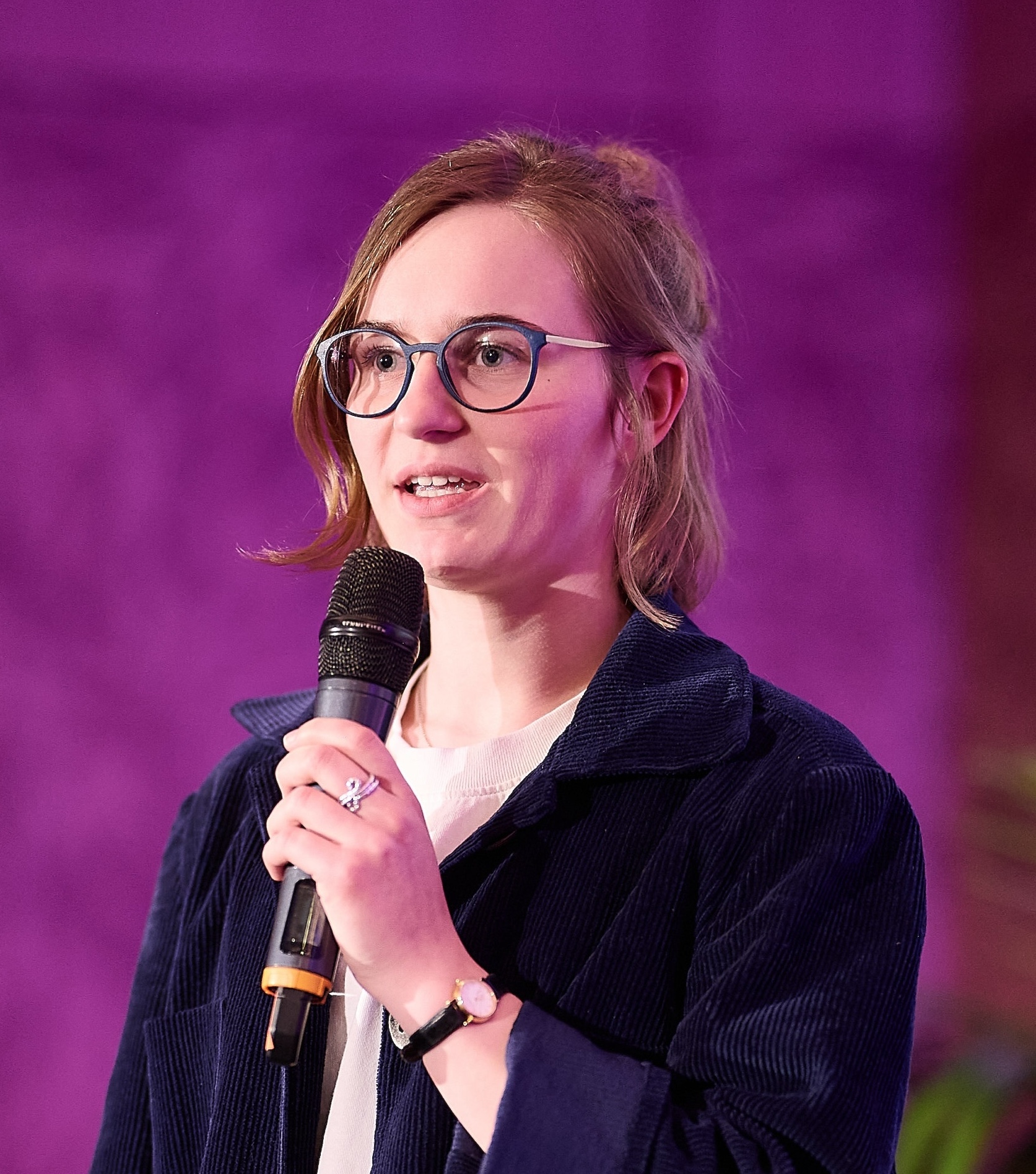 junge Frau spricht in Mikrofon, sie trägt eine blaue Brille und einen dunkelblauen Blaser, ihre Haare sind halb offen, der Hintergrund ist lila