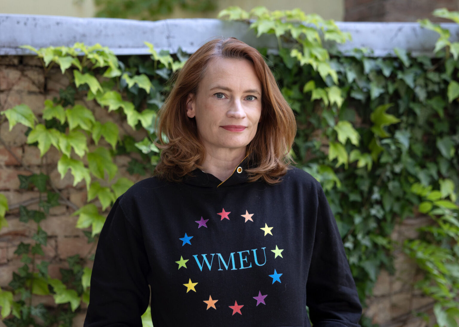 Claudia vor begrünter Wand mit einem Hoodie von Wikimedia Europe in Regenbogenfarben