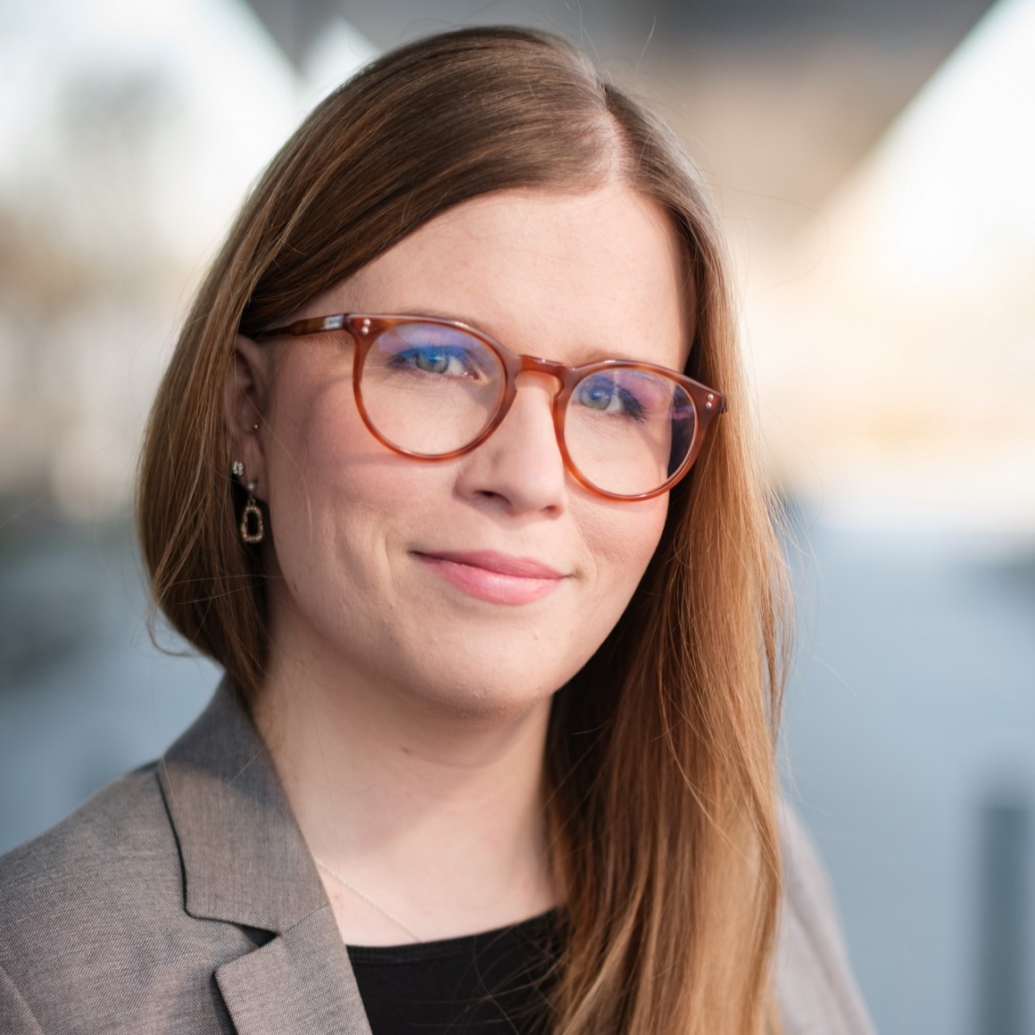 Frau mit langen, glatten rot-blonden Haaren und oranger Brille mit grauem Blazer