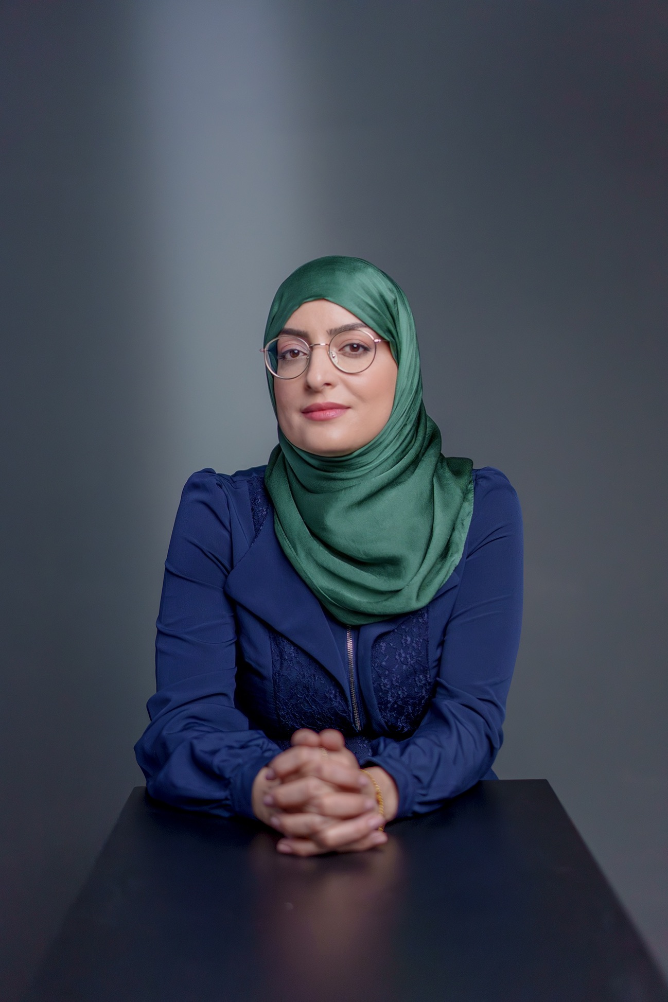 Porträt von Dr. Asmaa El Idrissi mit grünem Hijab und dunkelblauem Blazer. Sie sitzt mit gefalteten Händen an einem Tisch vor grauem Hintergrund und blickt in die Kamera.