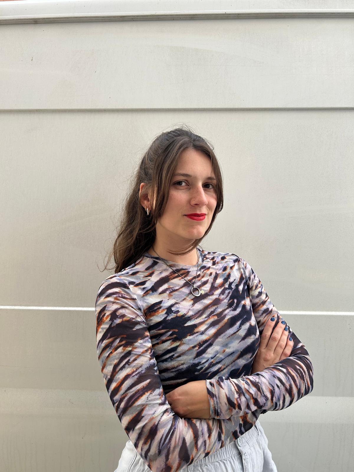 Kristina Kobl hat lange braune Haare und trägt ein bunt gemustertes Oberteil. Sie verschränkt die Arme und lächelt in die Kamera.