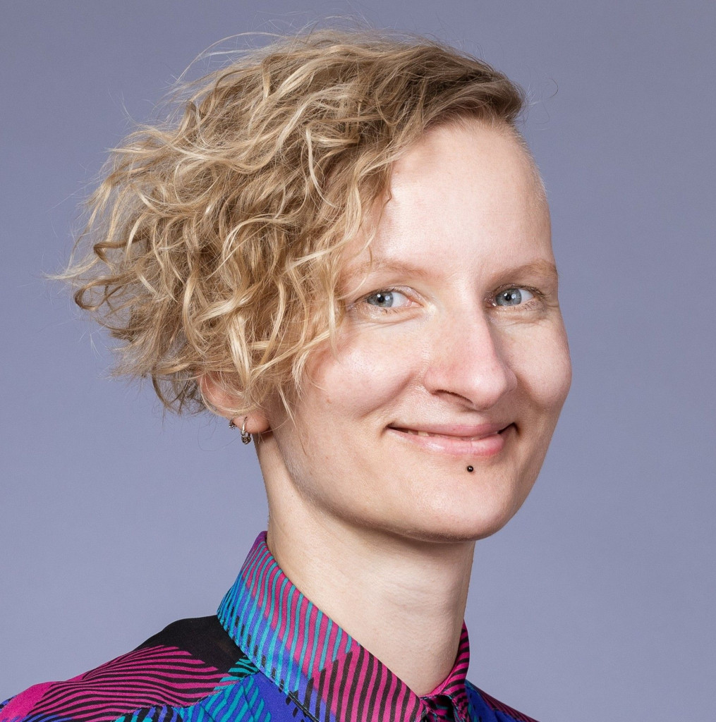 Porträtfoto einer lächelnden Person mit kurzen, blonden Locken und Undercut. Sie trägt ein bunt gemustertes Hemd in Pink, Türkis und Blau. Neutraler Studiohintergrund, Blick direkt in die Kamera.
