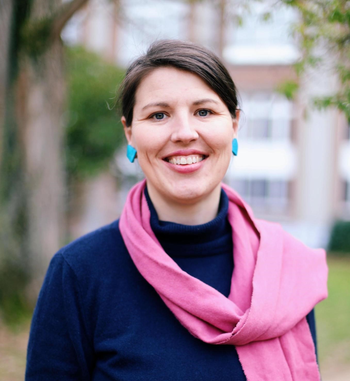 A picture of a female. I wear a blue roll neck with a pink scarf and blue hear rings. I look at the camera and smile.