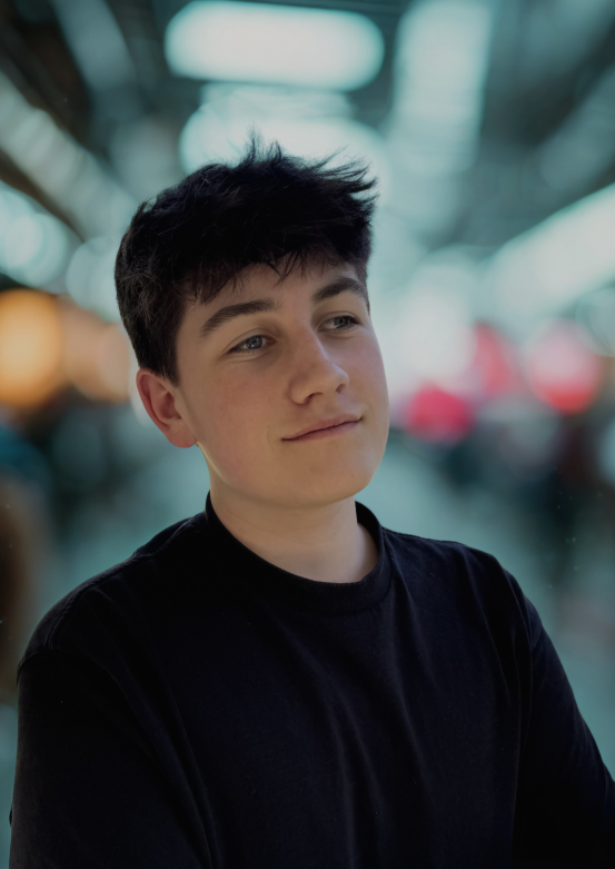Junger Mann in schwarzem T-Shirt mit dunklen, kurzen Haaren blickt zur Seite. Der Hintergrund ist stark verschwommen im urbanen Stil.