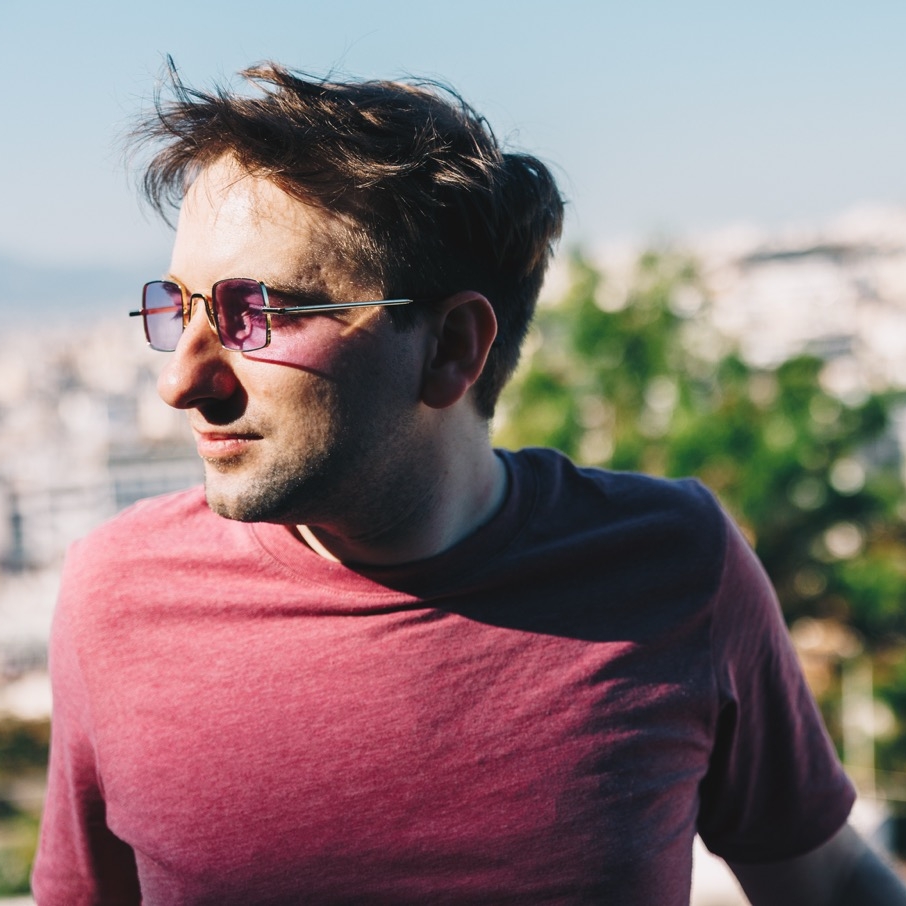 Ein Bild auf dem eest9 nach rechts blickt. eest9 wirkt nachdenklich aber zufrieden. Im Hintergrund sieht man verschwommen eine Stadt. eest9 trägt ein rotes T-Shirt und eine Violette quadratische Brille