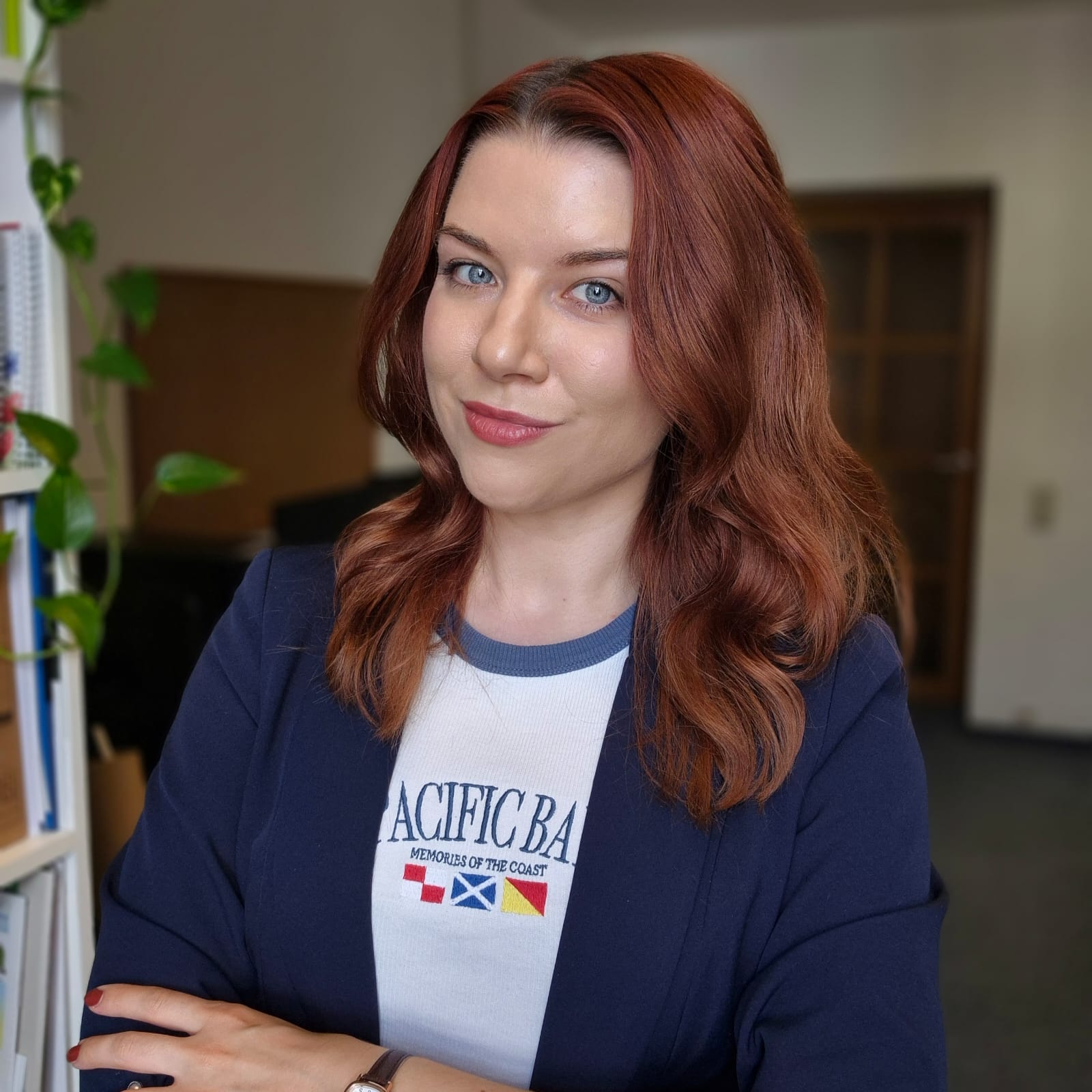 Eine junge Frau mit roten Haaren, dunkelblauem Blazer und einem weißen T-Shirt mit Aufschrift steht mit verschränkten Armen in einem Raum, der schemenhaft im Hintergrund erkennbar ist.