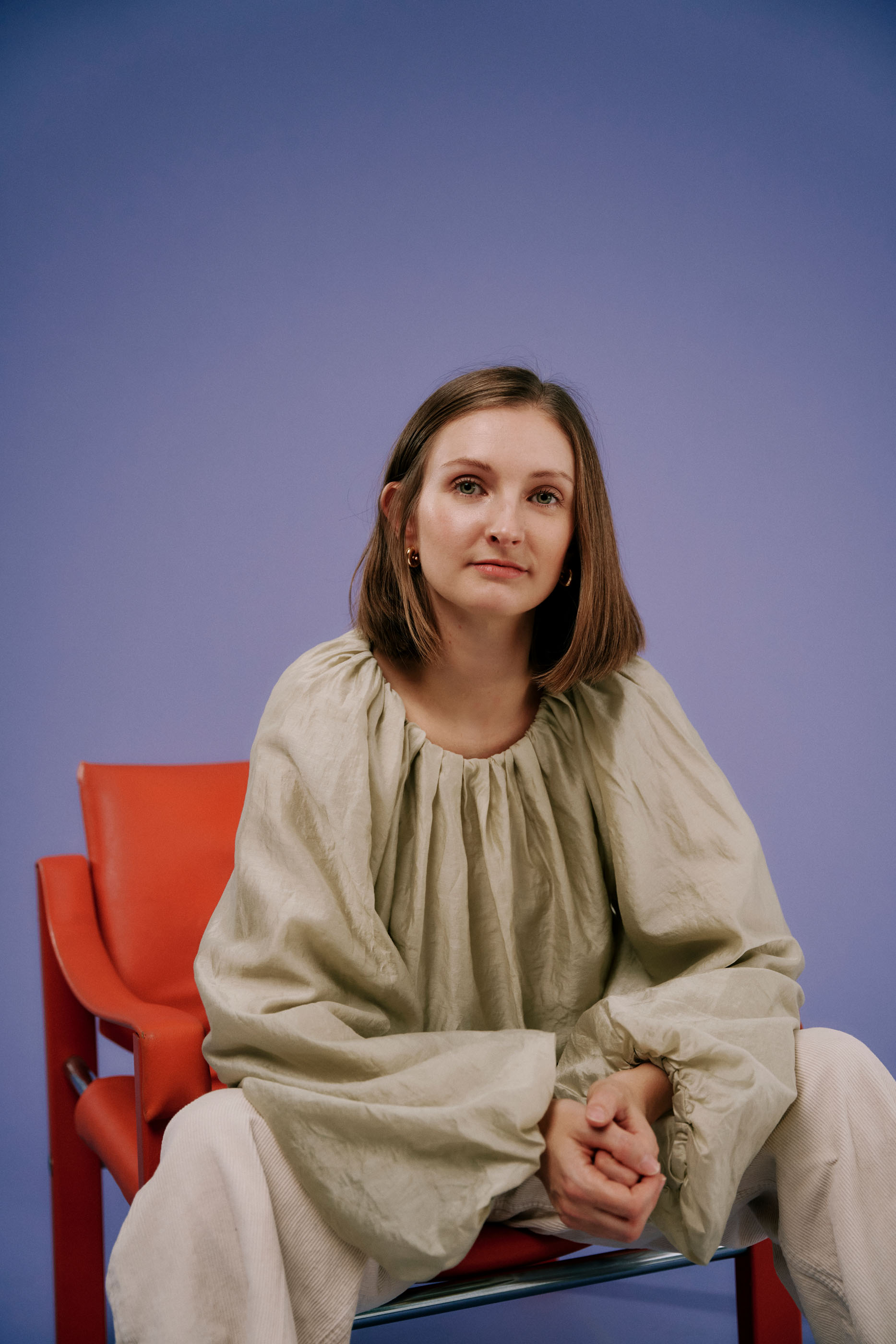 Jana Reininger da Rosa auf einem roten Stuhl vor lila Hintergrund. Sie verschränkt ihre Hände und sieht ernst in die Kamera.