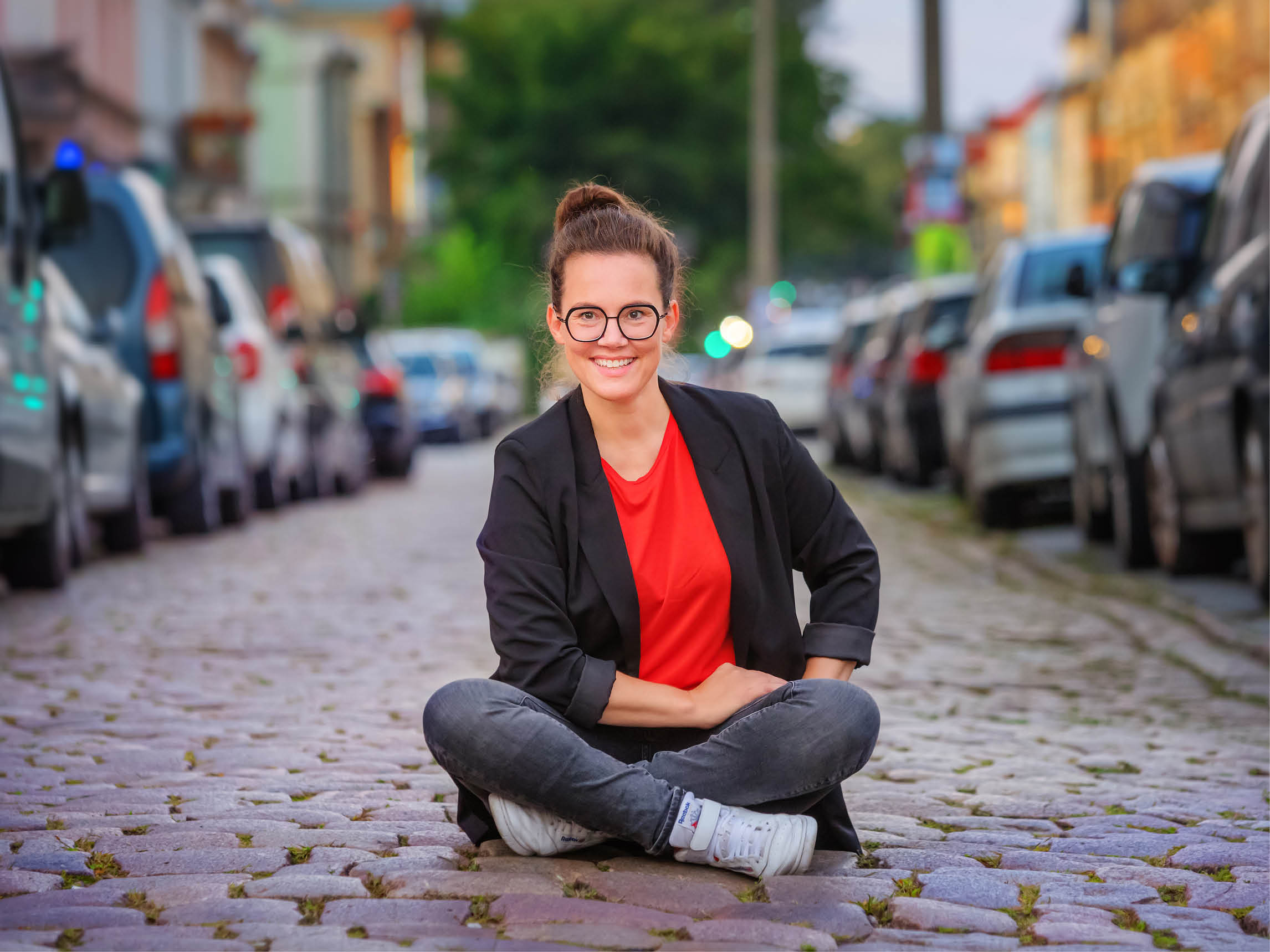 Katja sitzt im Schneidersitz auf einer Kopfsteinpflasterstraße. Um sie herum sehr viele geparkte Autos.