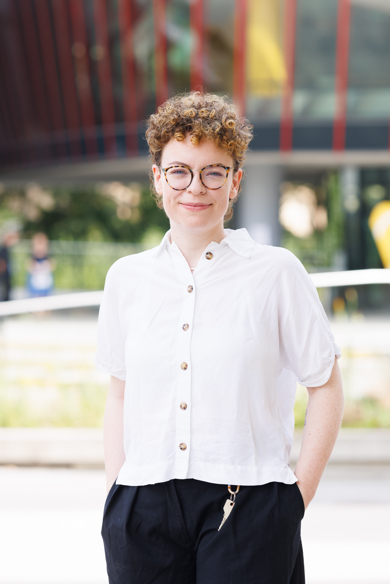 Foto von Person mit blond-braunen Locken, blasser Haut, mit weißer Bluse und schwarzer Hose vor einem Universitätsgebäude
