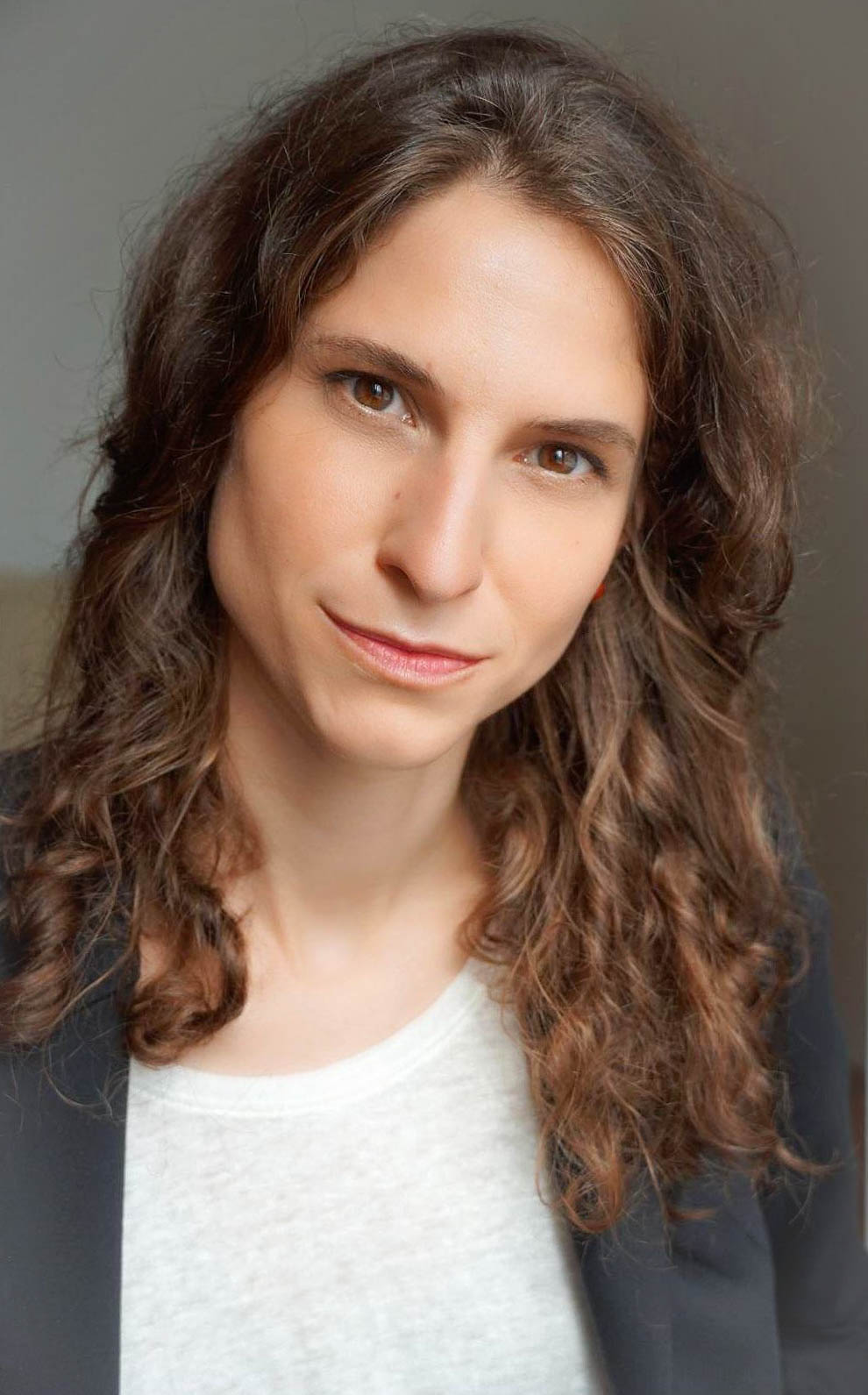 Professional headshot of Lia Musitz with long brown hair, wearing a black blazer over a white T-shirt, with a slight smile, against a light background.